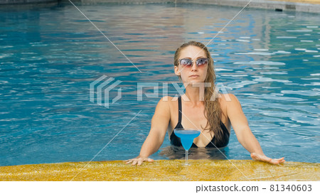 The wide middle shot pretty woman drinking blue cocktail alcohol liquor in swimming pool at hotel. Portrait of sexy girl in pool outdoor. 81340603