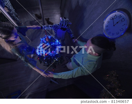 The family fast and fanny decorates the Christmas tree. Top view. Time on the clock is 23.45 on wall. Happy dad, mom, daughter are run around tree. The family fast and fanny decorates the Christmas tree. Top view. Time on the clock is 23.45 on wall. Happy dad, mom, daughter are run around tree. 81340657