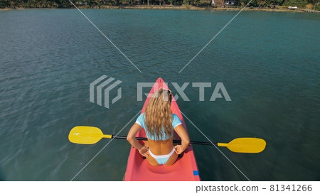 Seductive blonde woman in blue swimsuit sails on pink plastic kayak along azure sea at exotic resort backside upper above view. 81341266