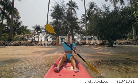 Young woman in sunglasses pulls pink plastic canoe with paddle to azure sea water from beach with palms. 81341381
