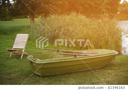 Wooden lounge chair and a boat on the grass of the shore of calm lake Wooden lounge chair and a boat on the grass of the shore of calm lake 81346163