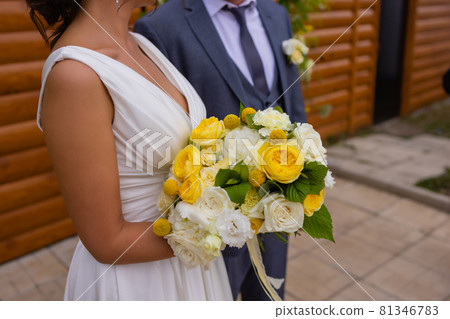 bride and groom standing on green grass and holding a bouquet of white and yellow flowers with green. 81346783