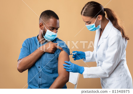 Young doctor in face mask making coronavirus vaccine injection to black male patient over beige studio background 81348975