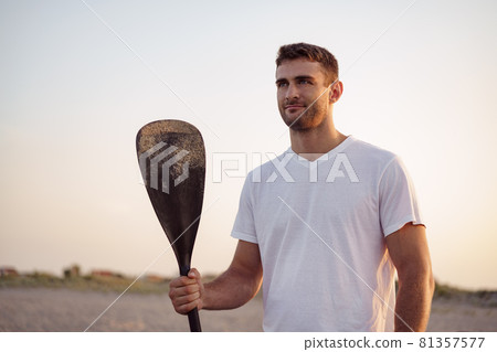 Young fit man standing on the beach and holding paddle Young fit man standing on the beach and holding paddle 81357577