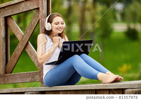 Woman with laptop sits outdoors in cordless headphones. Woman with laptop sits outdoors in cordless headphones. 81357978