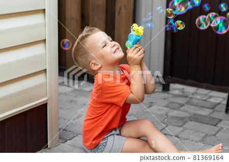 Portrait of cute little bond kid boy enjoy having fun play blowing soap bubbles siiting at home yard outdoors on bright warm summer day against house wall. Child healthy outside nature activities 81369164