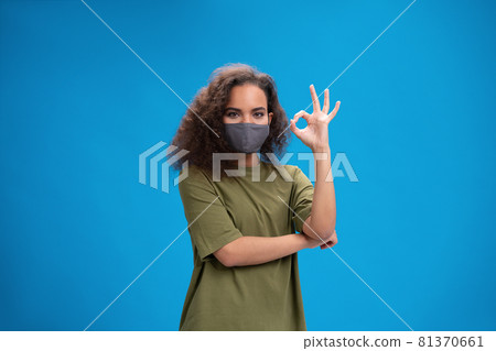 Young African American girl in khaki t-shirt in reusable face mask showing OK gesture, to prevent others from corona COVID-19 and SARS cov 2 infection isolated on pink background Young African American girl in khaki t-shirt in reusable face mask showing OK gesture, to prevent others from corona COVID-19 and SARS cov 2 infection isolated on pink background 81370661