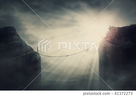 Person on the edge of precipice determined to overcome the obstacle by passing an old slackline bridge. The pursuit of success, danger and risk concept. Tightrope between two peaks, metaphoric scene Person on the edge of precipice determined to overcome the obstacle by passing an old slackline bridge. The pursuit of success, danger and risk concept. Tightrope between two peaks, metaphoric scene 81372273