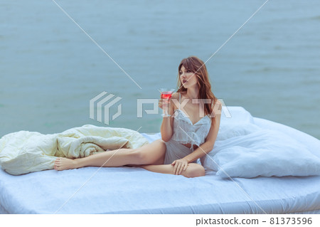 Woman with a glass of wine on a bed in the ocean. 81373596