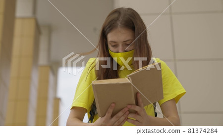 A schoolgirl girl in a mask reads a book while standing at the school, protection against coronavirus, covid-19 pandemic, air filtration through a mask filter, modern education for adolescents 81374284