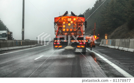 Orange highway maintenance gritter truck spreading de-icing salt, crystals dropping on the ice covered asphalt road during overcast day Orange highway maintenance gritter truck spreading de-icing salt, crystals dropping on the ice covered asphalt road during overcast day 81378977