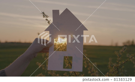 A man holds a paper house in his hand at sunset in the sky, the concept of a happy family, the construction and delivery of residential premises, the settlement of people, the sale and lending of a 81383129
