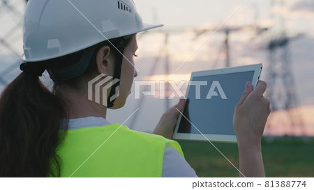 An adult woman electrician is working with a tablet on modern technologies next to a power plant, setting up energized stations through satellite control of networks, volt-installed towers, a gadget 81388774