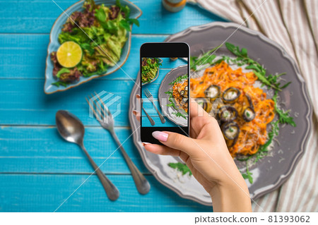 Photographing food concept - woman takes picture of pasta with eggplants, tomato, cheese, arugula and salad Photographing food concept - woman takes picture of pasta with eggplants, tomato, cheese, arugula and salad 81393062