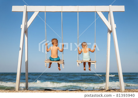 Back view couple of cute little adorable small caucasian sibling kids enjoy having fun swinging and playing at playground sea sand beach against blue ocean water wave and clear sky. Family vacation 81393940