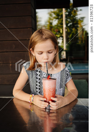 Funny little girl drinks through cocktail tube strawberry cocktail. family cafe, summer veranda Funny little girl drinks through cocktail tube strawberry cocktail. family cafe, summer veranda 81396728