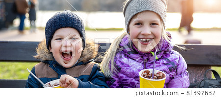 Two cute adorable siblings children sitting on bench drink delicious yummy hot chocolate, tea cocoa from paper cups during walk at city street park or backyard outdoors. Brother and sister enjoy fun 81397632