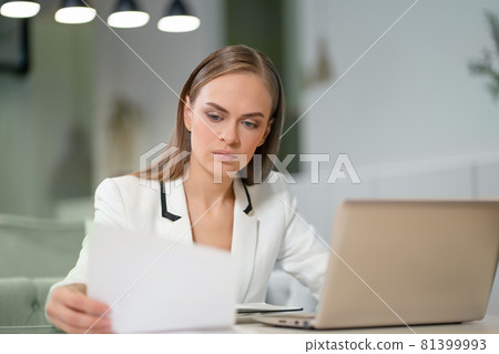 Portrait of a beautiful businesswoman in white jacket working with documents in her hands sitting in front of laptop. Office worker looking at her working place Portrait of a beautiful businesswoman in white jacket working with documents in her hands sitting in front of laptop. Office worker looking at her working place 81399993