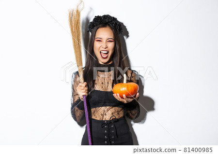 Image of beautiful asian woman dressed-up as a witch for halloween party, holding broom and pumpkin, standing over white background Image of beautiful asian woman dressed-up as a witch for halloween party, holding broom and pumpkin, standing over white background 81400985