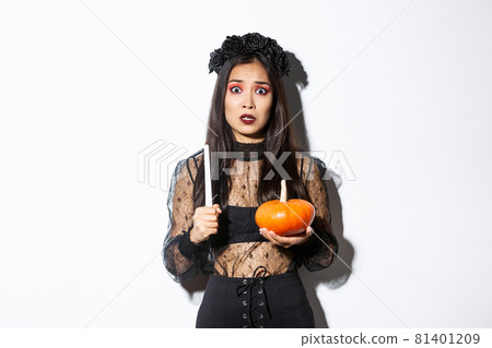 Image of asian woman panicking, being scared of witchcraft on halloween, standing in witch costume with candle and pumpkin 81401209