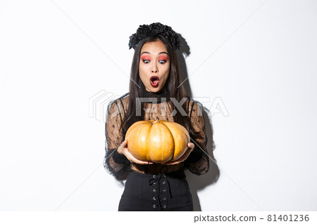 Image of amazed asian girl looking at big pumpkin fascinated, wearing gothic dress and black wreath, standing over white background Image of amazed asian girl looking at big pumpkin fascinated, wearing gothic dress and black wreath, standing over white background 81401236
