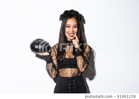 Sassy attractive asian woman in witch costume looking cunning and smiling, holding pumpking, going trick or treating on halloween, standing over white background Sassy attractive asian woman in witch costume looking cunning and smiling, holding pumpking, going trick or treating on halloween, standing over white background 81401238