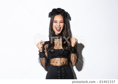 Cheerful asian woman in gothic lace dress and black wreath showing credit card and rejoicing, standing over white background 81401337