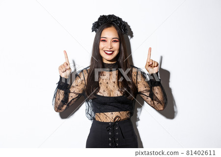 Cheerful elegant asian woman in black gothic dress and wreath, smiling happy and showing your logo, pointing fingers up at something about halloween, standing over white background Cheerful elegant asian woman in black gothic dress and wreath, smiling happy and showing your logo, pointing fingers up at something about halloween, standing over white background 81402611
