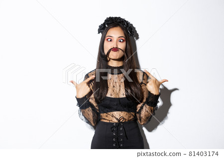 Portrait of funny and cute asian woman in witch costume making silly grimaces, pointing at herself, made moustache from hair strand Portrait of funny and cute asian woman in witch costume making silly grimaces, pointing at herself, made moustache from hair strand 81403174