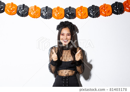 Image of happy and satisfied asian female in witch costume showing super, thumbs-up gesture and smiling pleased, standing against pumpkins banner 81403178