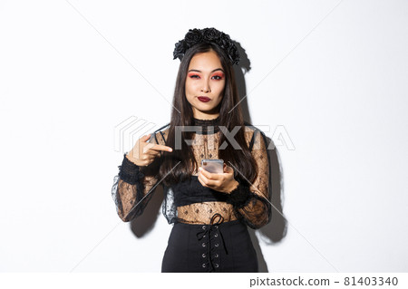 Sassy beautiful asian woman in gothic dress and black wreath pointing finger at mobile phone, showing something about halloween 81403340