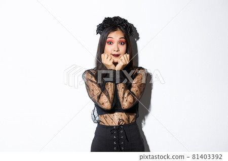Excited cute asian girl in witch costume looking hopeful at camera, daydreaming while standing over white background, wearing gothic lace dress with wreath 81403392