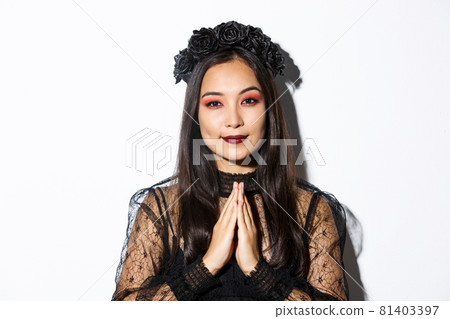 Close-up of young asian woman in black gothic dress and wreath holding hands in pray, girl wearing witch costume and celebrating halloween Close-up of young asian woman in black gothic dress and wreath holding hands in pray, girl wearing witch costume and celebrating halloween 81403397