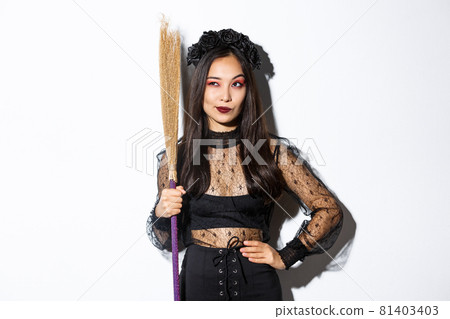 Portrait of smart thoughtful asian girl looking at upper left corner with pleased smirk, holding broom, wearing witch costume for halloween party, standing over white background 81403403