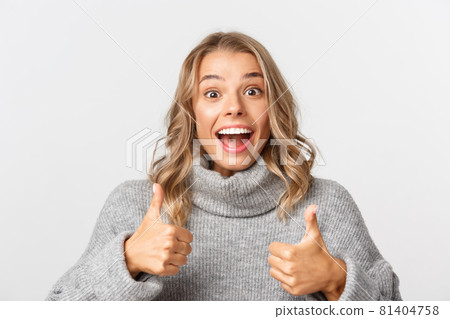 Close-up of cheerful blond girl in grey sweater, smiling and showing thumbs-up, like and approve something, standing over white background 81404758