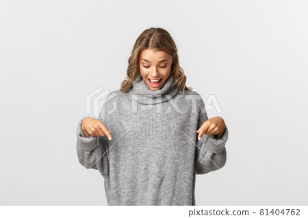 Cheerful blond girl rejoicing as looking and pointing fingers down at your logo banner, standing in grey sweater over white background 81404762