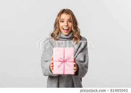 Beautiful young woman in grey sweater, giving gift, congratulating someone with birthday, standing over white background Beautiful young woman in grey sweater, giving gift, congratulating someone with birthday, standing over white background 81405043