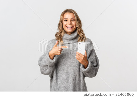 Cheerful blond girl with white smile pointing finger at mobile phone, showing something in internet, standing over white background 81405060