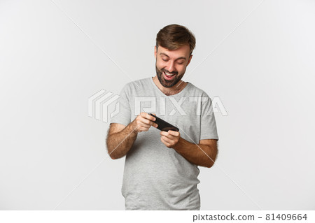 Image of adult bearded man in gray t-shirt, playing mobile games and having fun, standing over white background 81409664