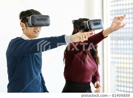 Portrait of cute smiling young Asian lover couple having fun with playing a new video game with virtual reality headset glasses together. Their posture like they try to touch screen in a virtual world Portrait of cute smiling young Asian lover couple having fun with playing a new video game with virtual reality headset glasses together. Their posture like they try to touch screen in a virtual world 81409889