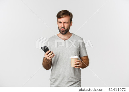 Image of disappointed frowning guy in gray t-shirt, drinking takeaway coffee and holding mobile phone, standing over white background Image of disappointed frowning guy in gray t-shirt, drinking takeaway coffee and holding mobile phone, standing over white background 81411972