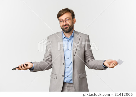 Image of clueless smiling businessman in glasses and gray suit, spread hands sideways and shrugging confused, holding mobile phone and credit card Image of clueless smiling businessman in glasses and gray suit, spread hands sideways and shrugging confused, holding mobile phone and credit card 81412006