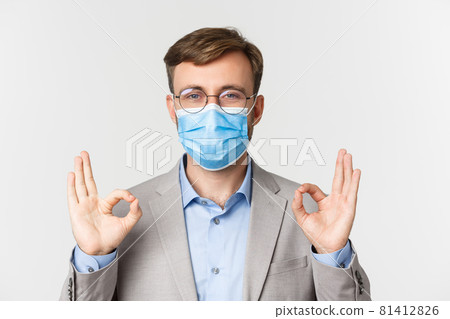 Concept of work, covid-19 and social distancing. Close-up of businessman in grey suit and medical mask, showing okay signs in approval, standing over white background 81412826