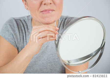 Caucasian woman looks in the mirror and removes hair on her chin herself with the help of tweezers. Hirsutism. Caucasian woman looks in the mirror and removes hair on her chin herself with the help of tweezers. Hirsutism. 81413811