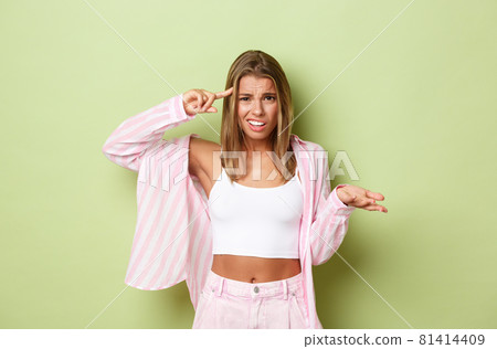Image of annoyed and frustrated blond woman in pink outfit, pointing at head and shrugging, scolding person for acting stupid or crazy, standing over green background 81414409