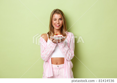 Portrait of beautiful glamour girl with blond hair, looking pleased, holding piece of cake and licking lips, standing over green background 81414536