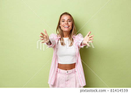 Image of friendly attractive girl with blond hair, wearing pink stylish costume, reaching hands forward to hold something or give a hug, standing over green background 81414539