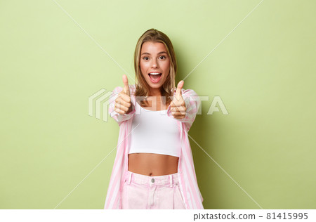 Image of attractive happy female model with blond hair, wearing pink shirt and jeans, showing thumbs-up in approval, like something, standing over green background 81415995