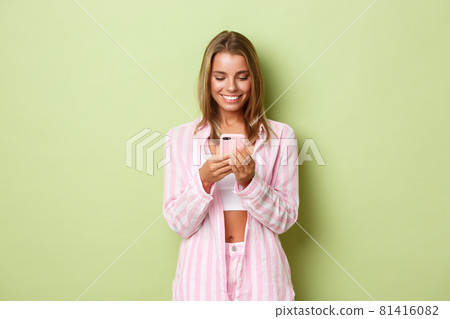 Image of beautiful blond woman in pink shirt, looking at mobile phone and smiling, standing over green background 81416082