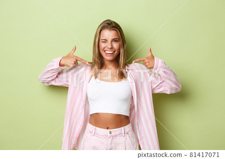 Portrait of sassy beautiful blond woman with perfect white smile, pointing fingers at logo on the center, showing something, standing over green background Portrait of sassy beautiful blond woman with perfect white smile, pointing fingers at logo on the center, showing something, standing over green background 81417071